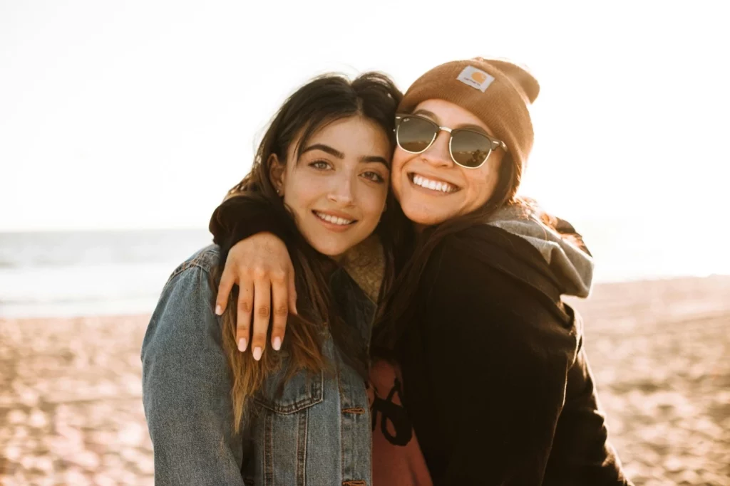 Two people hugging on a sandy beach with the ocean in the background during sunset. One in denim jacket, and the other is in a black hoodie and sunglasses.