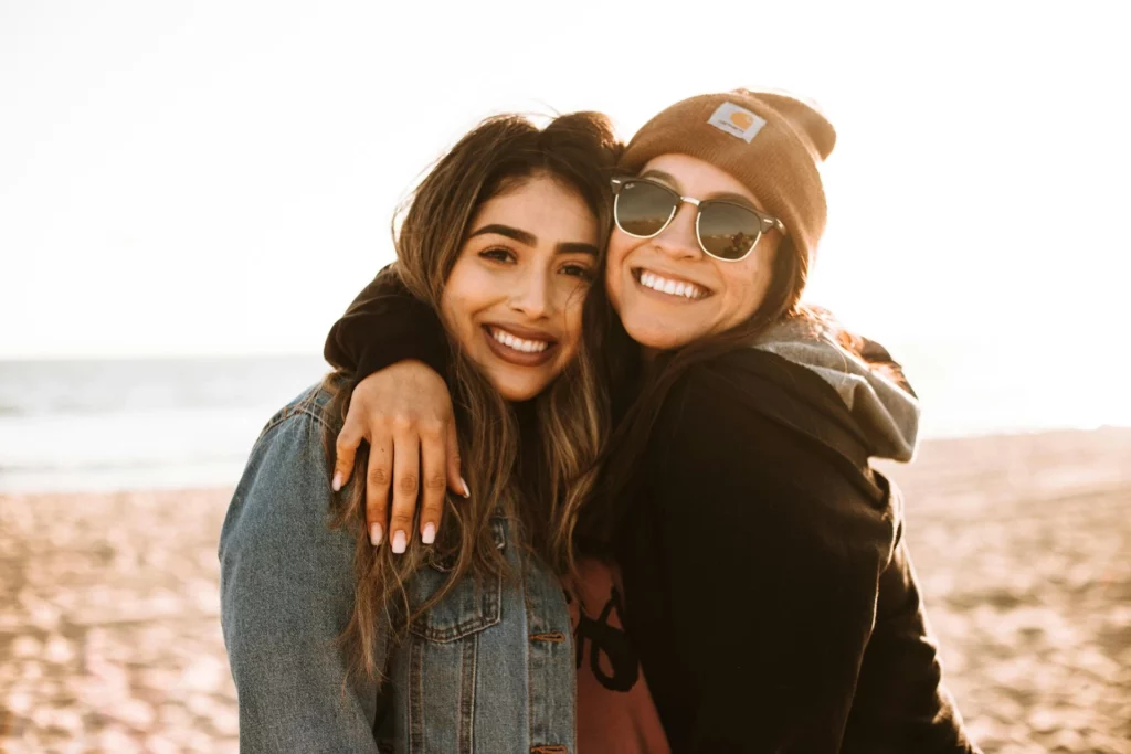 Two people hugging on a sandy beach with the ocean in the background during sunset. One wears a denim jacket, and the other is in a black hoodie with a brown beanie featuring a Carhartt logo.